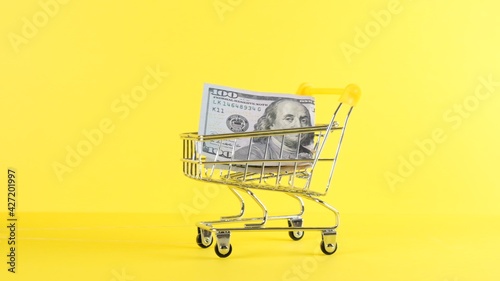 Shopping cart and one hundred dollar bill on a yellow background, closeup. Black Friday Shopping and Discount Concept