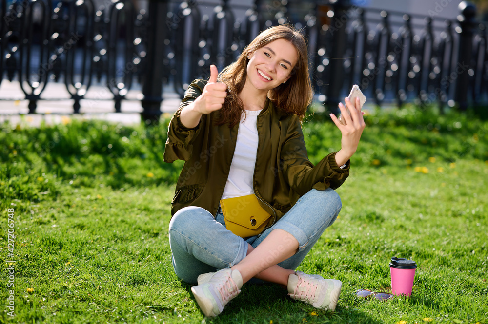 Smiling attractive young woman relaxes in public park sitting on the grass uses smartphone and shows thumb up gesture