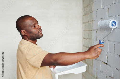 Young African man in beige t-shirt holding paintroller while standing in front of brick wall of living-room and painting it into white color