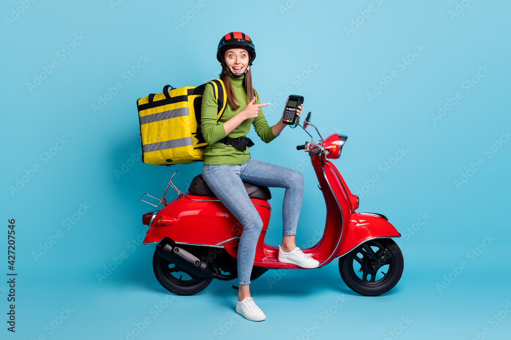 Full length photo portrait of girl pointing finger terminal big yellow package on red scooter isolated on pastel blue colored background Full length photo portrait of girl pointing finger terminal big yellow package on red scooter isolated on pastel blue colored background