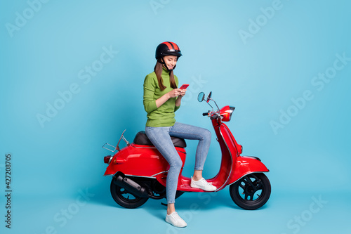 Full length photo portrait of woman holding phone in two hands sitting on red scooter isolated on pastel blue colored background
