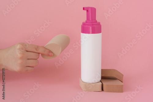 Facial cleansing sponges in a woman's hand with cosmetic bottle, face soap, foam, on pink background