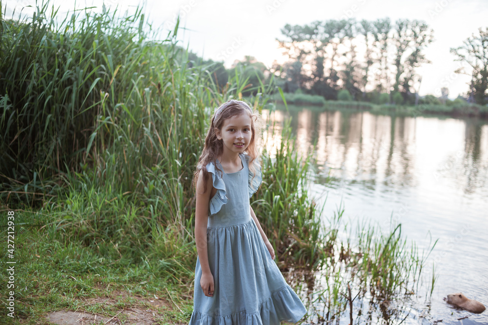 A girl in a long blue dress stands on the shore of the lake near nutria in the water. Child watching with interest on coypu swimming. Young lady and Myocastor coypus in public park in summer. A girl in a long blue dress stands on the shore of the lake near nutria in the water. Child watching with interest on coypu swimming. Young lady and Myocastor coypus in public park in summer.