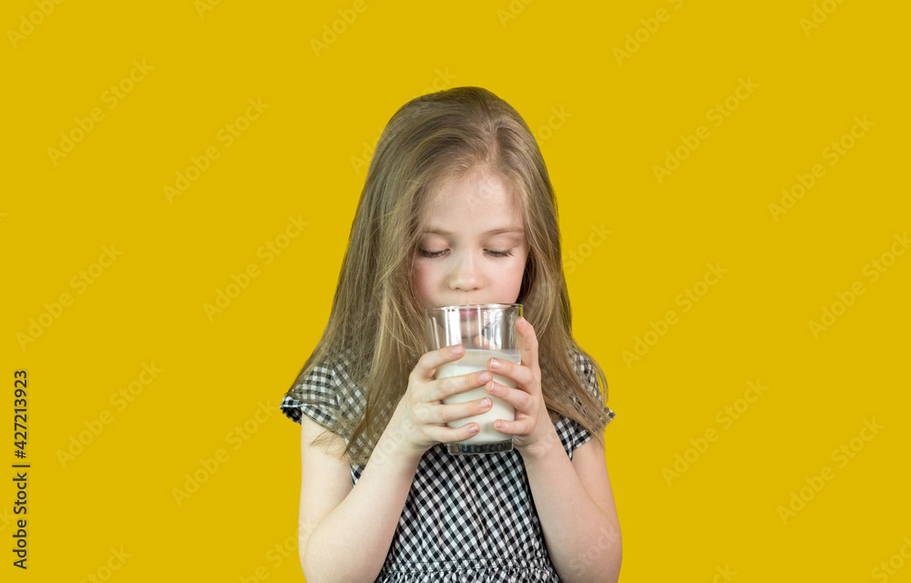 A cute little girl with blonde hair holds and drinks milk from a glass. It stands on a yellow background.Milk is good for children. She is healthy and happy. There is a place for the text.Soft focus. A cute little girl with blonde hair holds and drinks milk from a glass. It stands on a yellow background.Milk is good for children. She is healthy and happy. There is a place for the text.Soft focus.