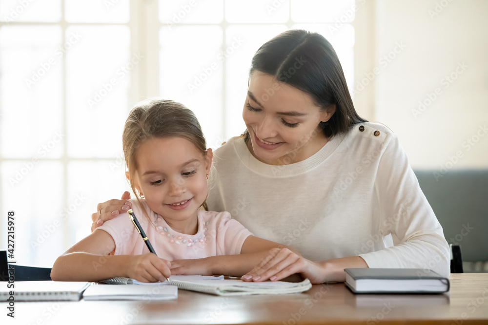 Happy young Caucasian mom and small daughter handwrite in exercise book prepare school home task together. Loving caring mother help little girl child with homework. Distant education concept. Happy young Caucasian mom and small daughter handwrite in exercise book prepare school home task together. Loving caring mother help little girl child with homework. Distant education concept.