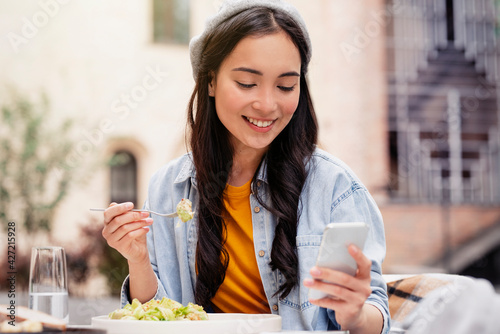 A beautiful young Asian woman is using an application to send a instant message in her smartphone device while eating a salad at the restaurant 