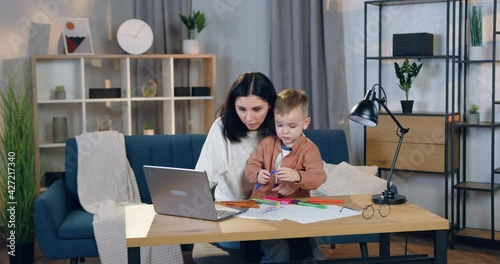 Good-looking loving confident woman is distracted from work on laptop and helping to draw her small curious son photo