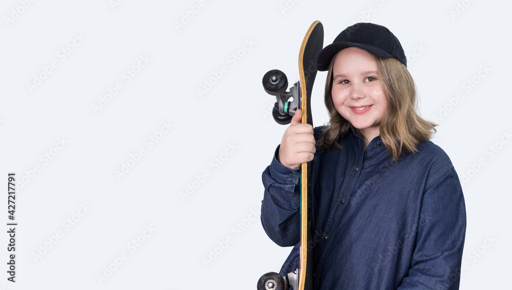 a teenage girl stands in casual clothes, wearing a cap and holding a skateboard on a light background. She is content and happy. Skateboarding is a sport, a game, and a competition. Copy space. a teenage girl stands in casual clothes, wearing a cap and holding a skateboard on a light background. She is content and happy. Skateboarding is a sport, a game, and a competition. Copy space.