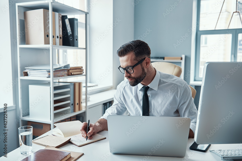 Concentrated young man in shirt and tie writing something down while sitting in the office Concentrated young man in shirt and tie writing something down while sitting in the office