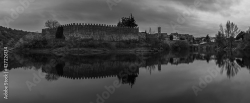 panoramic view of a wall and church tower in black and white reflected in a lake with cloudy sky