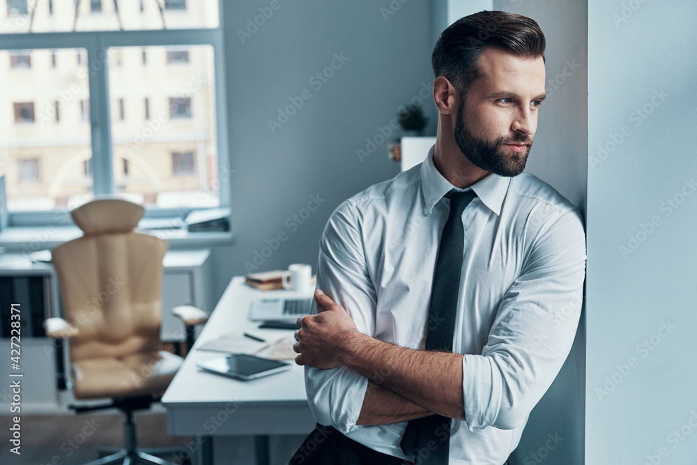 Thoughtful young man in formalwear looking away and keeping arms crossed while standing in office Thoughtful young man in formalwear looking away and keeping arms crossed while standing in office