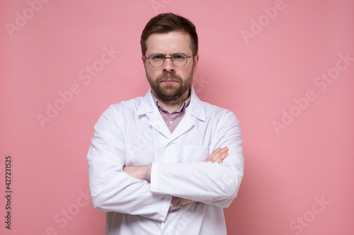 Serious doctor in a white coat and glasses looks sternly and stands with crossed arms. Pink background.
