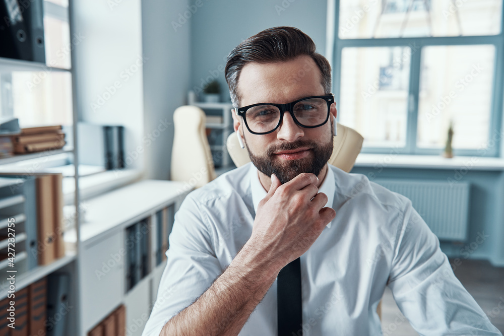 Handsome young man in formalwear looking at camera and keeping hand on chin while sitting in the office Handsome young man in formalwear looking at camera and keeping hand on chin while sitting in the office