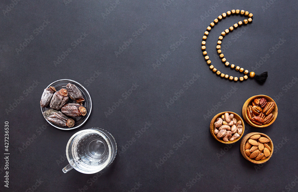 Ramadan Kareem and iftar muslim food and glass of clear water, modern holiday concept. Wooden bowls with nuts and dried fruits and the moon made of beads for rosary with tassel on a black background 