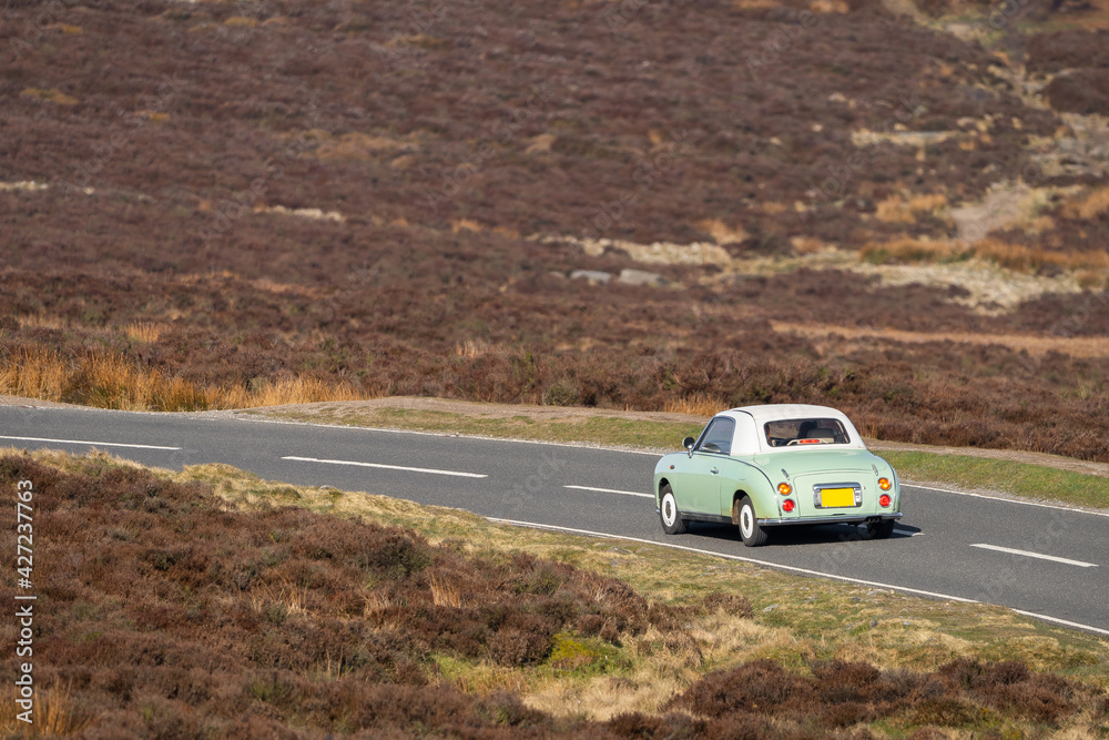 Fototapeta premium Old fashioned green classic car driving alone along narrow country road in Derbyshire peak district countryside moorland on sunny day. Picturesque summer scene tarmac lane winding through fields.