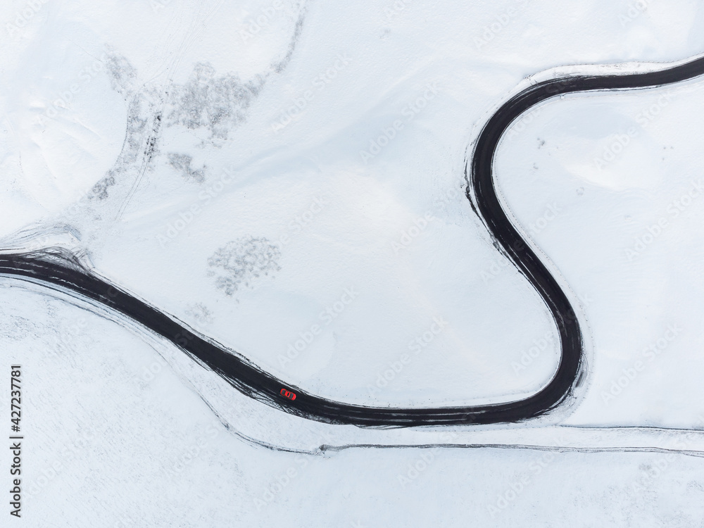 Aerial view of long winding black road through mountain range snow covered landscape. Looking down on single red car driving in icy conditions all alone winter alpine conditions. Drone shot of curves.