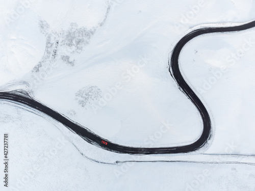 Aerial view of long winding black road through mountain range snow covered landscape. Looking down on single red car driving in icy conditions all alone winter alpine conditions. Drone shot of curves. © Matthew