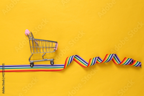 Empty shopping cart with rainbow ribbon on yellow background. Lgbt community. Shopping concept