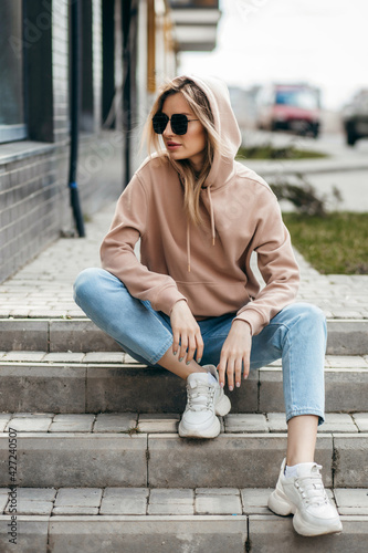 Fasion blonde woman in brown oversize hoodie, glasses and blue jeans posing sitting on the steps.  Mockup for logo or branding design © andrew_shots