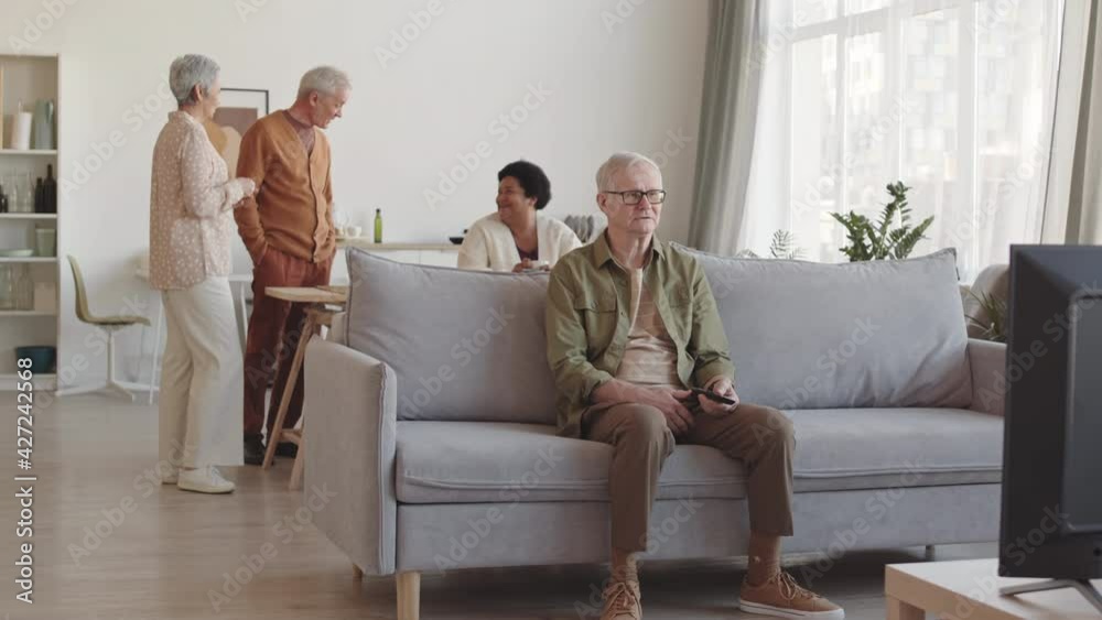 Wide shot of senior Caucasian man sitting on couch in living room, watching TV emotionally, calling for his friends they joining from background Wide shot of senior Caucasian man sitting on couch in living room, watching TV emotionally, calling for his friends they joining from background