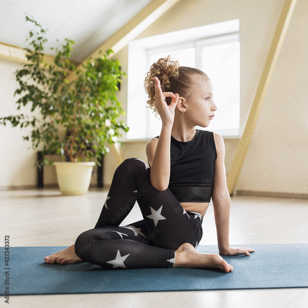 Obraz premium A little girl practicing yoga performs the Ardha Matsyendrasana, fish lord pose, sitting on a mat in the studio
