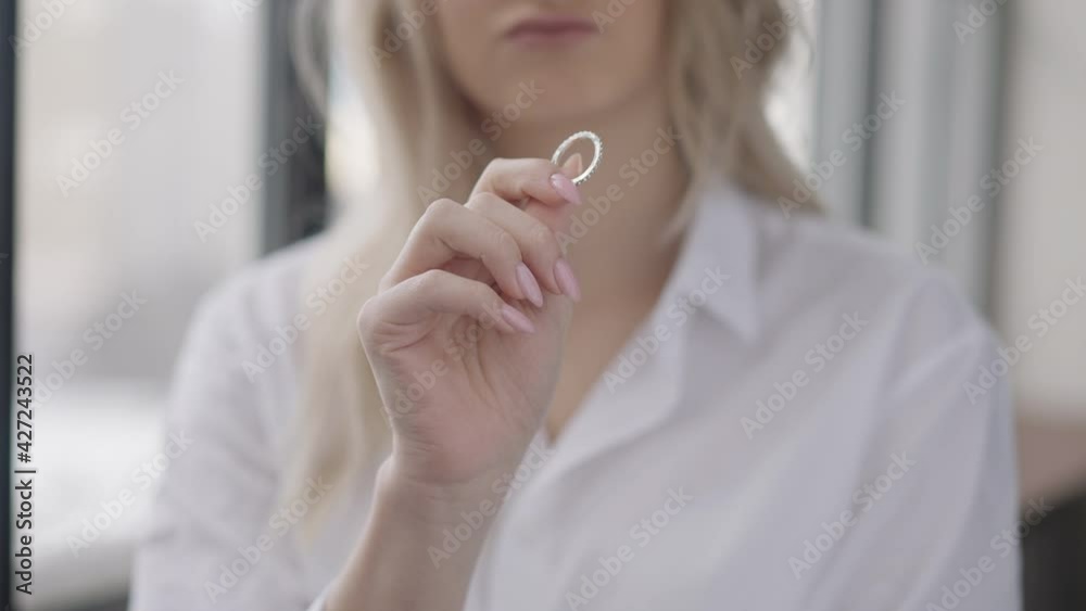 Close-up of ring in female Caucasian hand with blurred unrecognizable young woman sighing at background. Stressed cheated slim bride looking at jewelry thinking indoors. Heartbreak and divorce