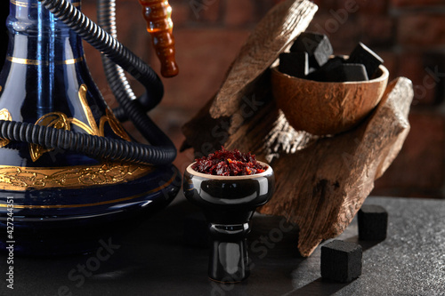 Photography Black glossy hookah bowl filled with apple shisha and prepared for smoking stands on a dark surface next to a beautiful hookah, charcoal and textured wood