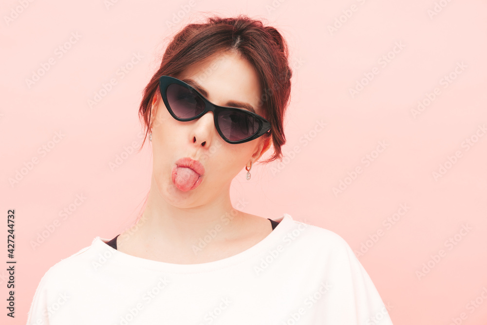 Young beautiful smiling female in trendy summer hipster clothes. Sexy carefree woman posing near pink wall in studio. Positive model having fun indoors. Cheerful and happy.Shows tongue in sunglasses