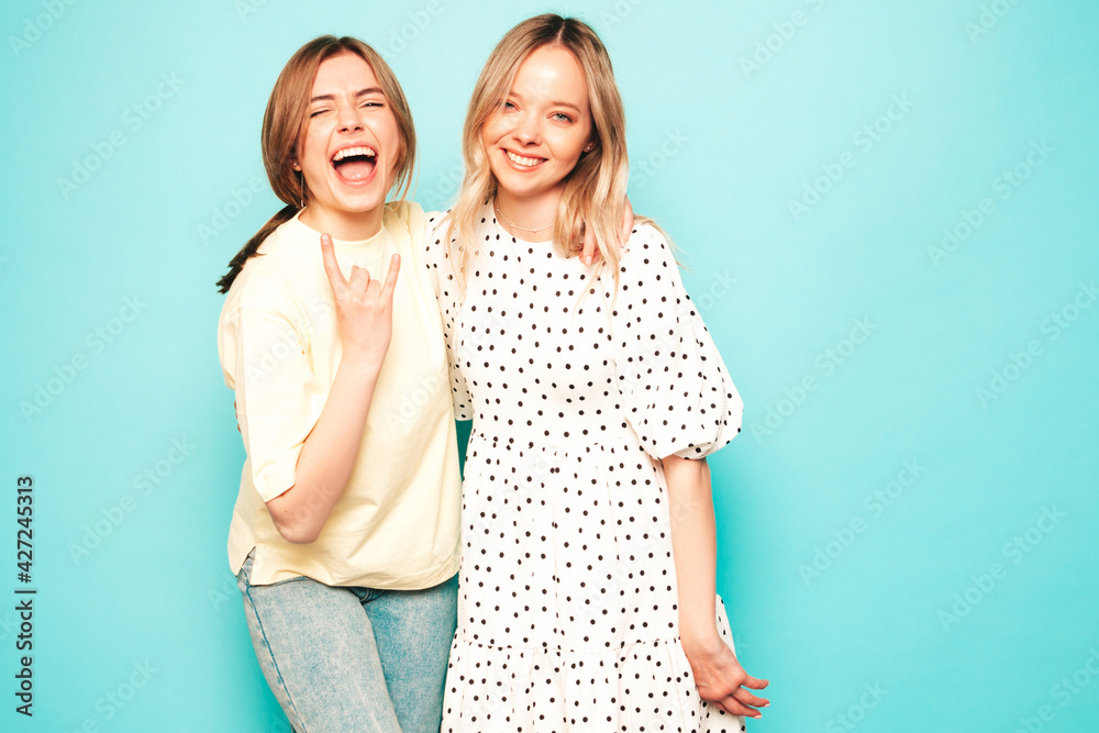Two young beautiful blond smiling hipster female in trendy summer clothes. Sexy carefree women posing near blue wall in studio. Trendy and positive models having fun Two young beautiful blond smiling hipster female in trendy summer clothes. Sexy carefree women posing near blue wall in studio. Trendy and positive models having fun