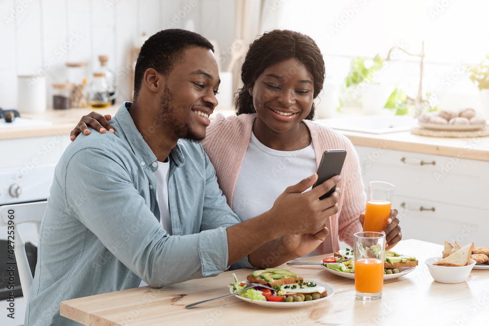 Happy African American Spouses Using Smartphone While Having Breakfast In Kitchen
