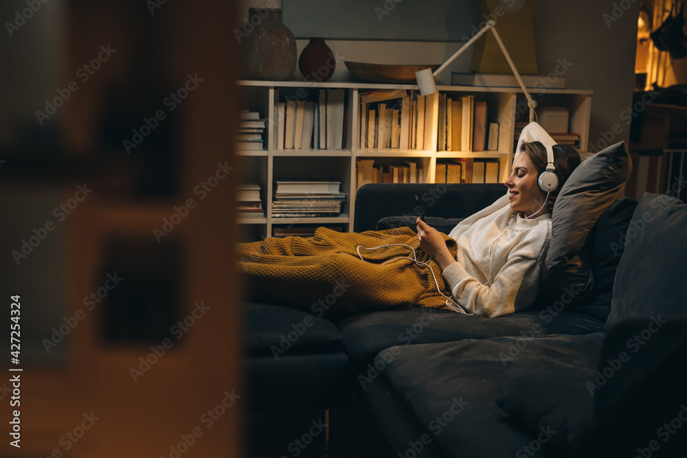 young woman listening to a music on headset. she is relaxing on sofa at her living room. evening scene