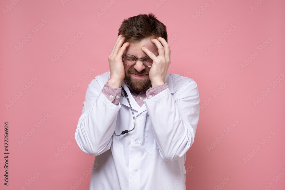 Doctor with a stethoscope around neck is stressed or has a severe headache, he holds head with his hands and makes an unhappy grimace. Doctor with a stethoscope around neck is stressed or has a severe headache, he holds head with his hands and makes an unhappy grimace.