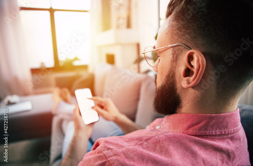 Young handsome happy bearded man in glasses is using smartphone for surfing in net, typing, and chatting, playing games, or working with some apps while relaxing on the couch at home