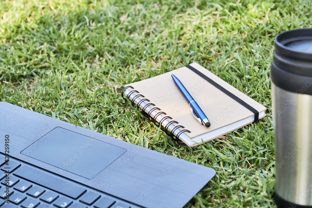 A laptop, a notebook with a pen and a thermal cup on the grass, outdoors A laptop, a notebook with a pen and a thermal cup on the grass, outdoors