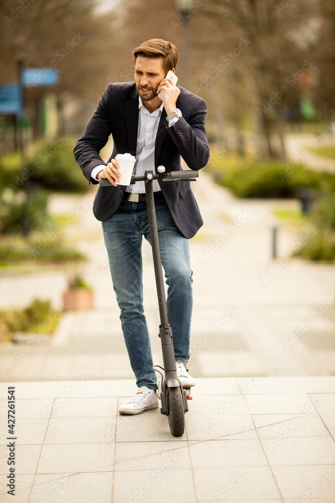 Young businessman using mobile phone while holding take away coffee cup on electric scooter