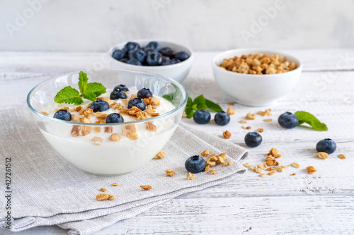 White yogurt granola in glass bowl with fresh berries and mint on white wooden table
