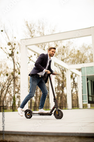 Young business man in a casual clothes riding an electric scooter by an office building on a business meeting