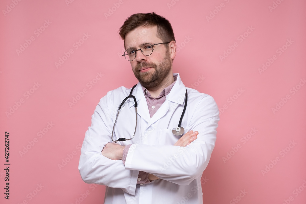 Calm, cute young doctor with a stethoscope around neck looks confidently into the camera with crossed arms. Pink background. Calm, cute young doctor with a stethoscope around neck looks confidently into the camera with crossed arms. Pink background.