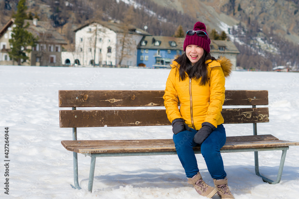 unique Winter holidays trip to amazing snow valley - young happy and beautiful Asian woman sitting on bench alone enjoying snowy mountain view - obrazy, fototapety, plakaty 