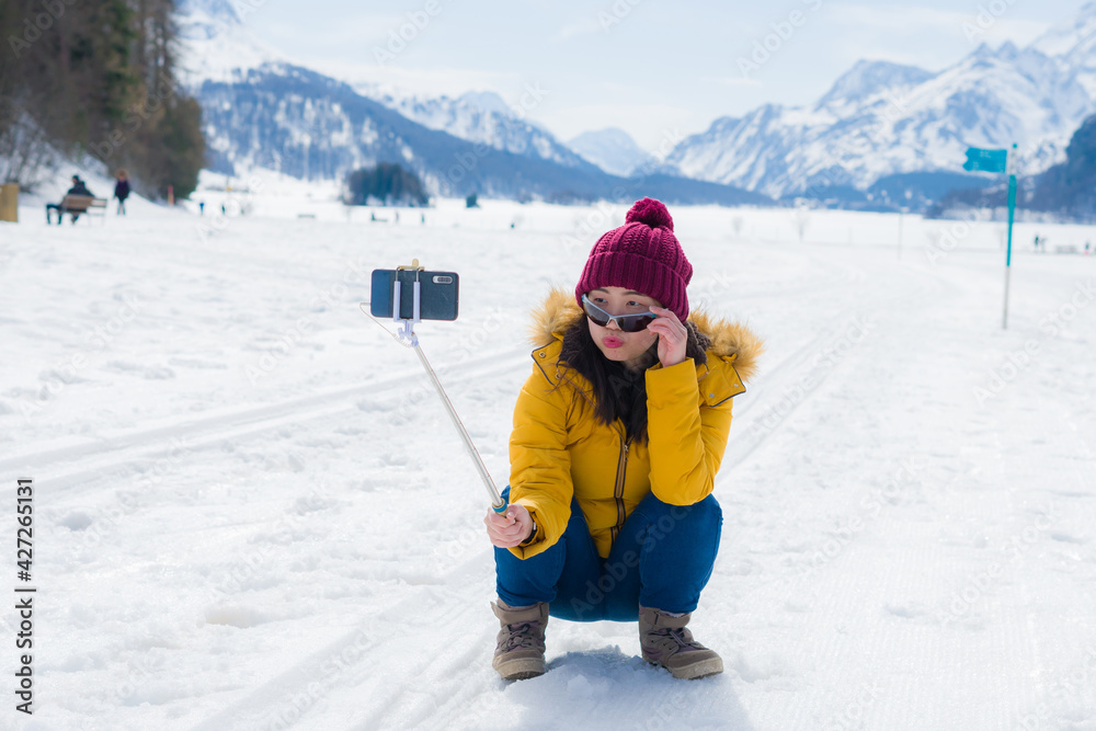 Winter holidays selfie in the snow - young happy and cheerful Asian Korean woman on taking self portrait with mobile phone in cold snowy mountain at Swiss Alps