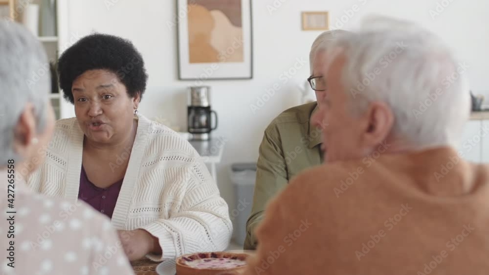 Medium close-up over shoulders of mature African woman and Caucasian man sitting at table at home, talking to unrecognizable friends over tea Medium close-up over shoulders of mature African woman and Caucasian man sitting at table at home, talking to unrecognizable friends over tea