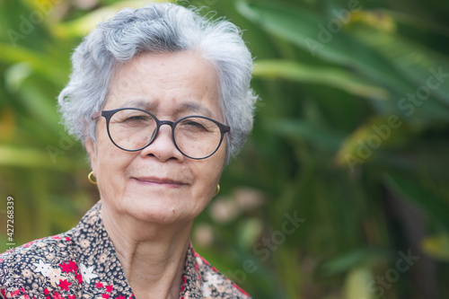 Portrait of a beautiful senior woman with short gray hair, wearing glasses, smiling and looking at the camera while standing in a garden. Space for text. Concept of aged people and relaxation