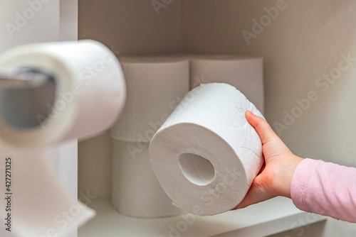 Roll of white toilet paper in hand. The child's hand takes a roll of toilet paper from the cabinet. Lots of toilet paper on background