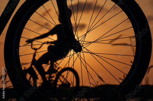Close-up silhouette of a bike wheel at sunset. The sun shines through the wheel of a bicycle with blurred bicycle rider figure
