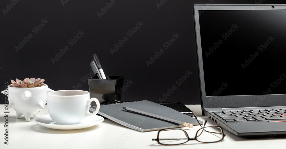 Home workspace for work or study in Office interior. Black laptop on white table black background. Glasses, cup of coffee, office supplies. Long web banner Home workspace for work or study in Office interior. Black laptop on white table black background. Glasses, cup of coffee, office supplies. Long web banner