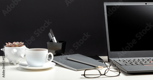 Home workspace for work or study in Office interior. Black laptop on white table black background. Glasses, cup of coffee, office supplies. Long web banner