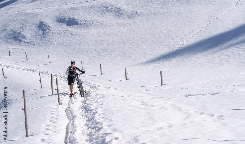 senior woman mountainbiking and pushing her e-mountainbike through a snow spot in early spring, in the Allgaeu Area, a part of the bavarian alps,Germany