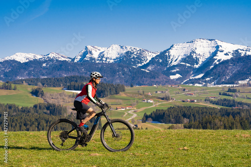 senior woman mountainbiking below the Nagelfluh mountain chain with Hochgrat summit on a e-mountainbike in early spring, in the Allgaeu Area near Steibis and Obers, a part of the bavarian alps,Germany