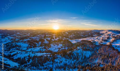 Picturesque winter panorama of mountain hills covered with snow and fir trees on a sunny clear day with the sun and blue sky. Pristine Nature Beauty Concept. Copyspace