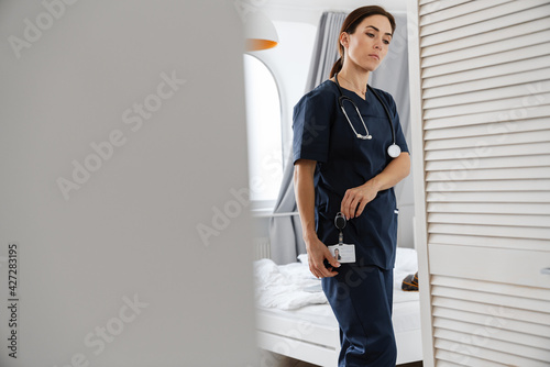 Focused mid woman doctor in medical suit fastening badge at home